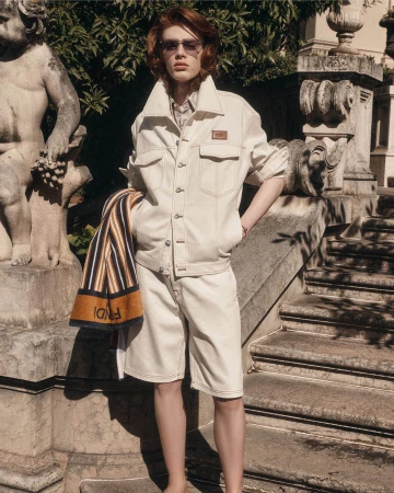 Model wearing a cream denim jacket and matching shorts standing outdoors on stone steps near carved classical statues.