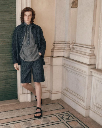 Model standing against classical architectural stone pillars wearing dark patterned jacket shorts and sandals