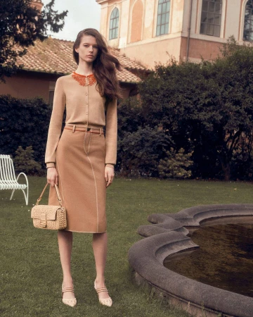 Model Achol Kuir in a camel cardigan and matching skirt holding a woven handbag on a lush lawn for Fendi.