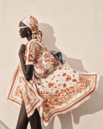Model Achol Kuir wearing matching patterned silk scarf and dress with a shoulder bag against a textured wall.