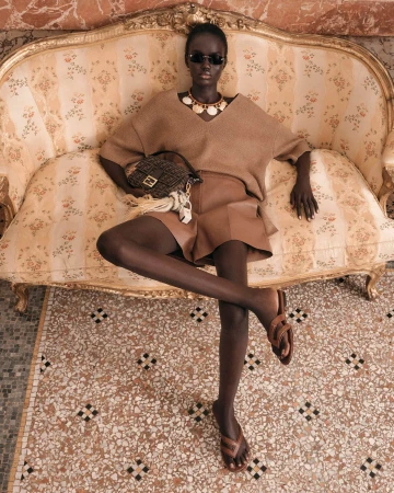 Model Achol Kuir seated on an ornate vintage sofa wearing a brown knit top, brown shorts, and sunglasses for Fendi