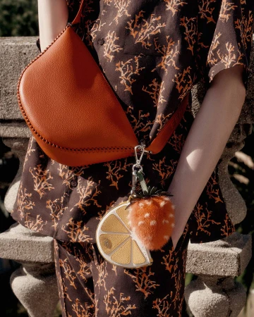 Model wearing a brown patterned short-sleeve shirt holding an orange leather shoulder bag with fruit charms attached