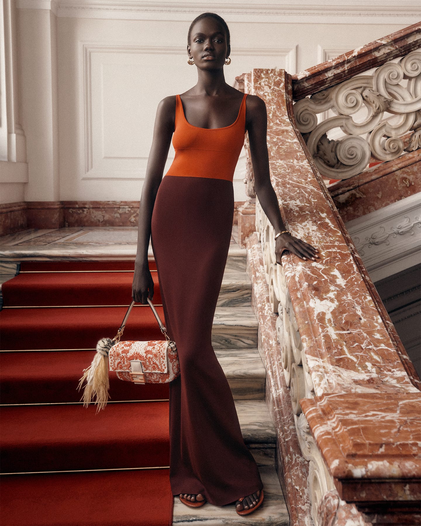 Model Achol Kuir in an orange and brown knit dress holding a patterned handbag on grand stairs for Fendi.