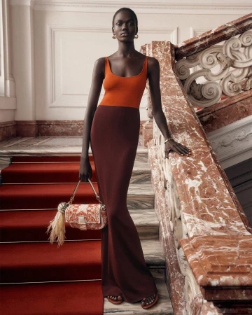 Model Achol Kuir in an orange and brown knit dress holding a patterned handbag on grand stairs for Fendi.