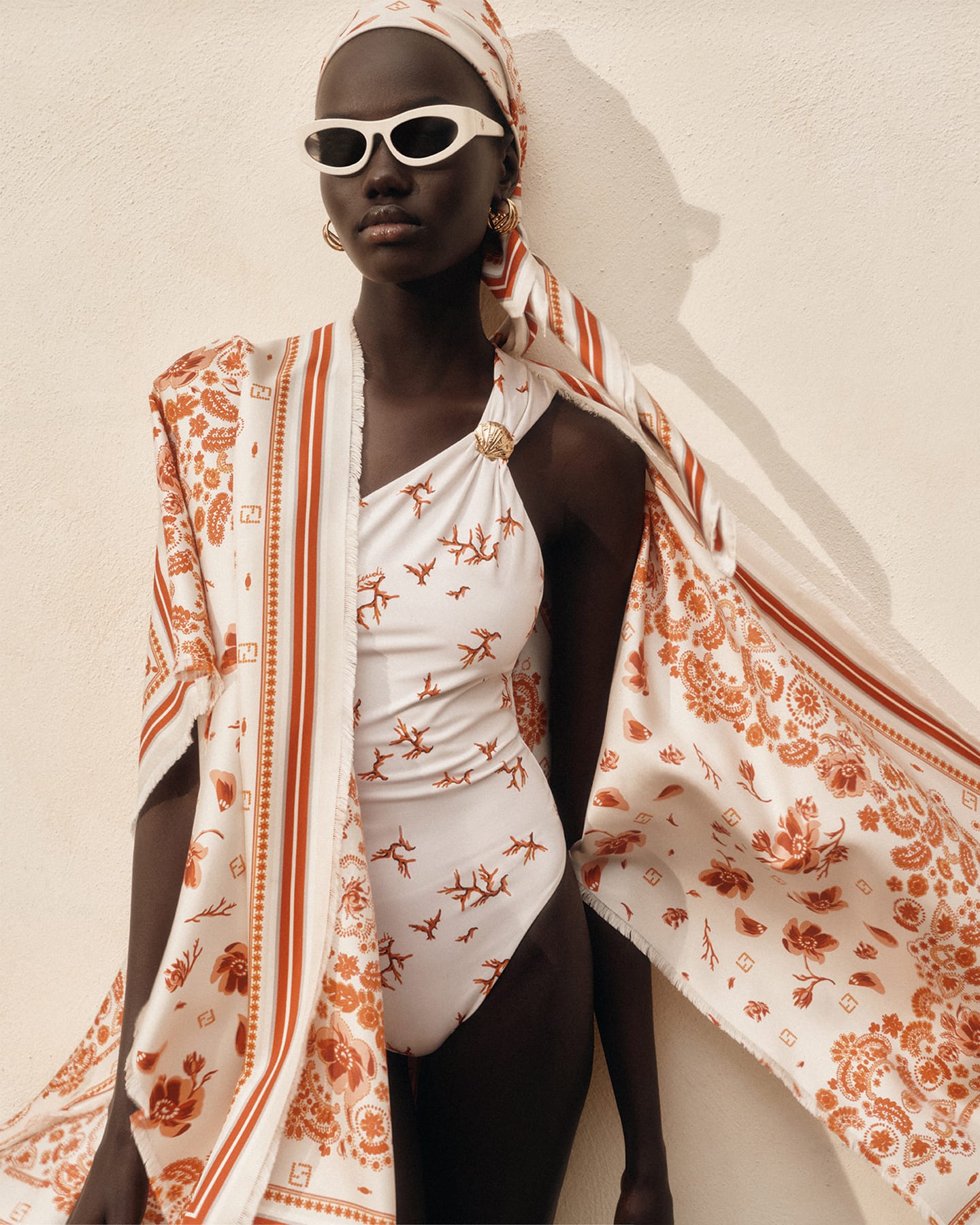 Model Achol Kuir wearing a patterned white swimsuit, matching silk scarf, and sunglasses against a plain neutral wall.
