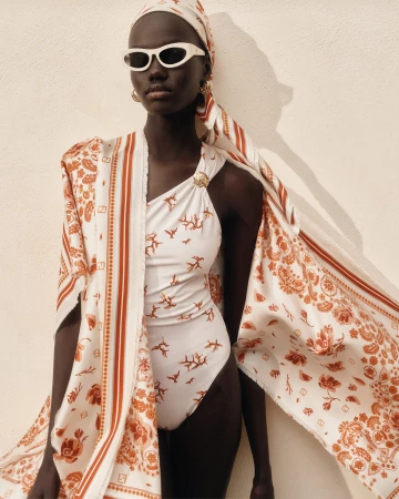 Model Achol Kuir wearing a patterned white swimsuit, matching silk scarf, and sunglasses against a plain neutral wall.