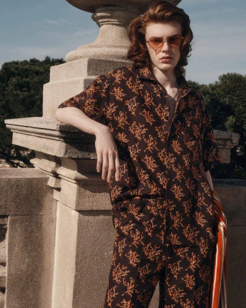 Model wearing a patterned short-sleeve shirt and matching trousers, leaning against a stone balustrade outdoors.