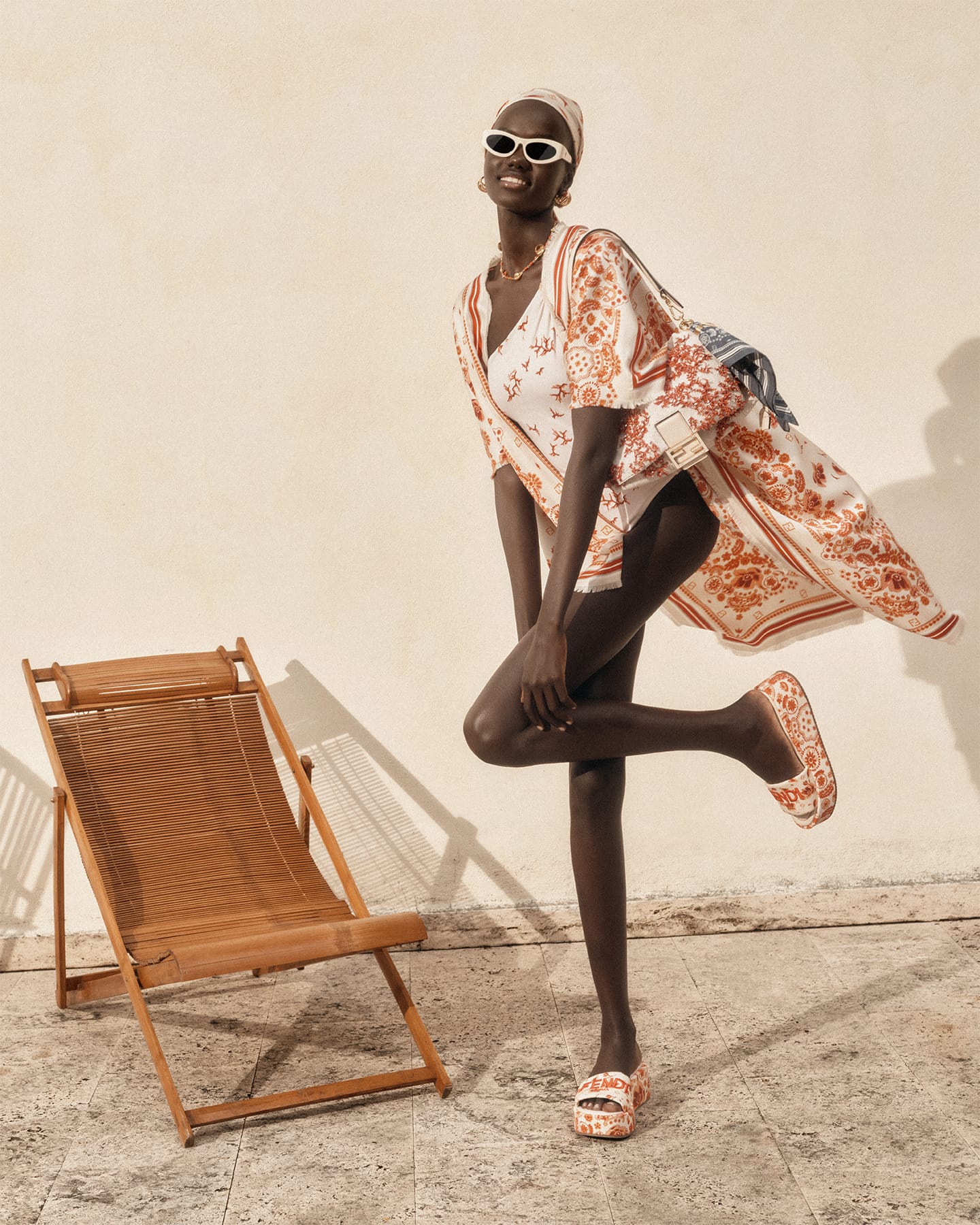 Model Achol Kuir in patterned apparel and white sunglasses posing next to a wooden deck chair against a plain wall.