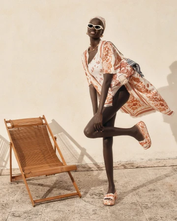 Model Achol Kuir in patterned apparel and white sunglasses posing next to a wooden deck chair against a plain wall.