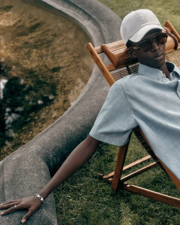 Model Achol Kuir relaxing in a wooden lounge chair outdoors wearing a light blue shirt and baseball cap