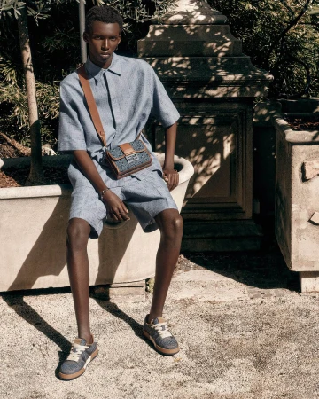Model Achol Kuir seated outdoors wearing a matching blue short-sleeve shirt and shorts with a brown crossbody bag for Fendi.