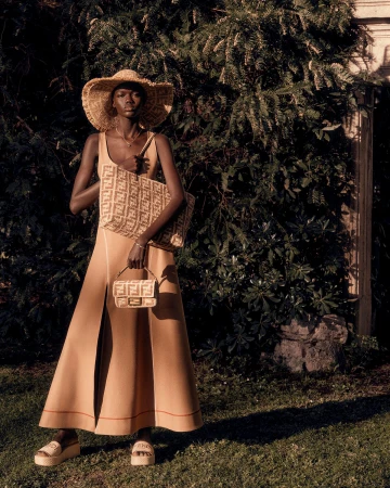 Model Achol Kuir wearing a straw sun hat, tan maxi dress, and platform sandals while holding monogrammed Fendi bags outdoors