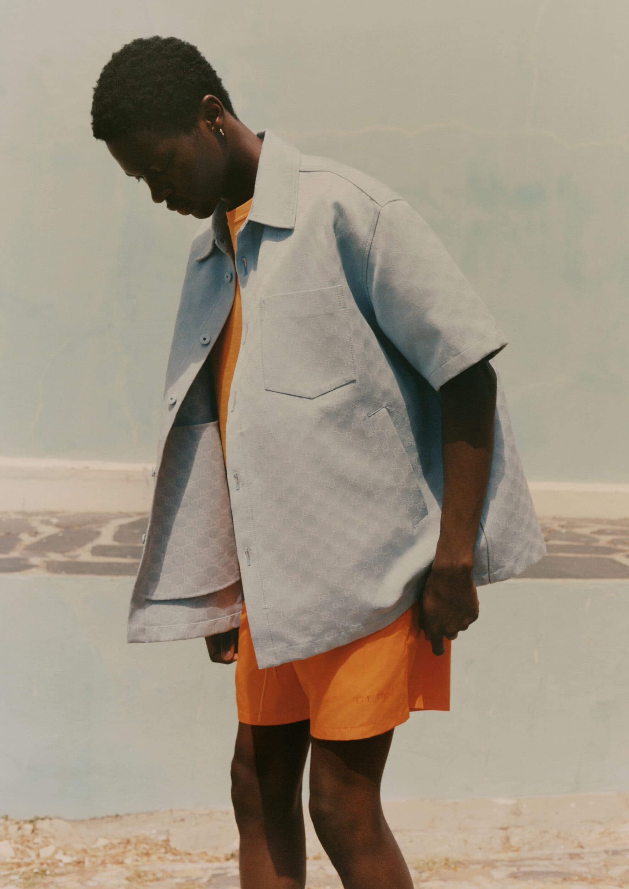 Model wearing a light blue short-sleeved button-up shirt and bright orange shorts against an outdoor background