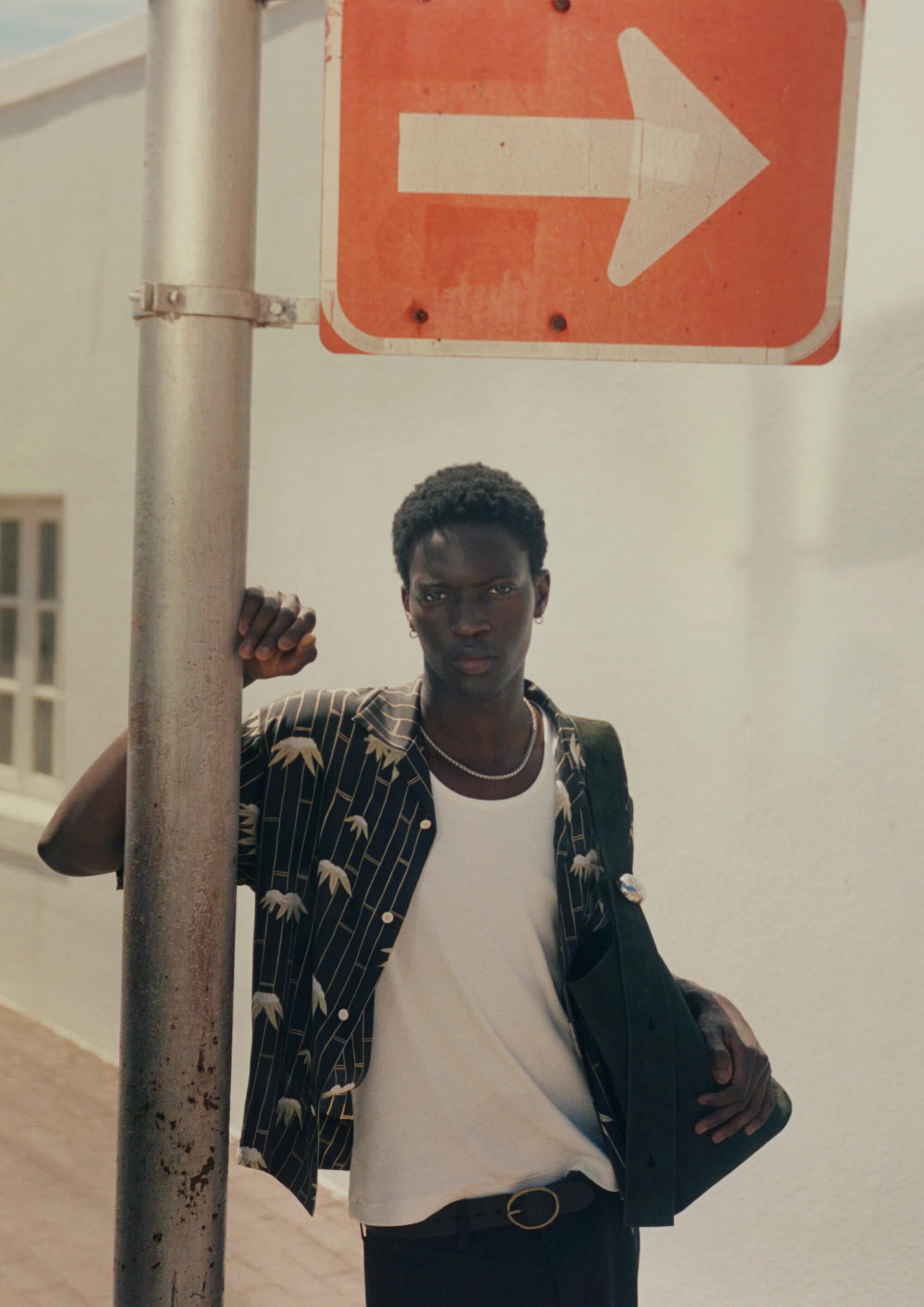 Model leaning against a metal pole beneath an orange directional arrow sign wearing a patterned shirt and white tank top.