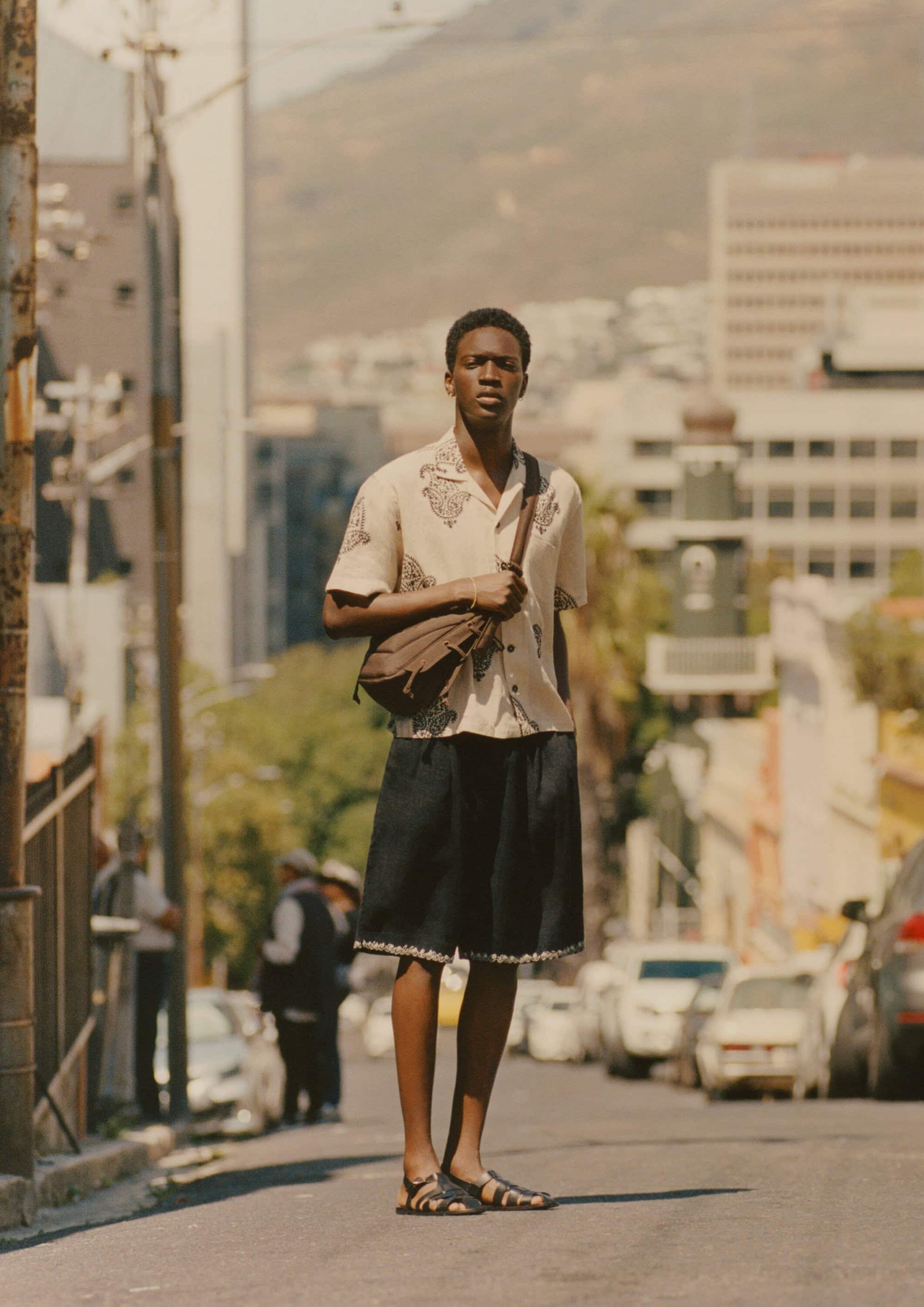 Man wearing a patterned shirt and dark shorts standing on a city street with parked cars and buildings in the background.