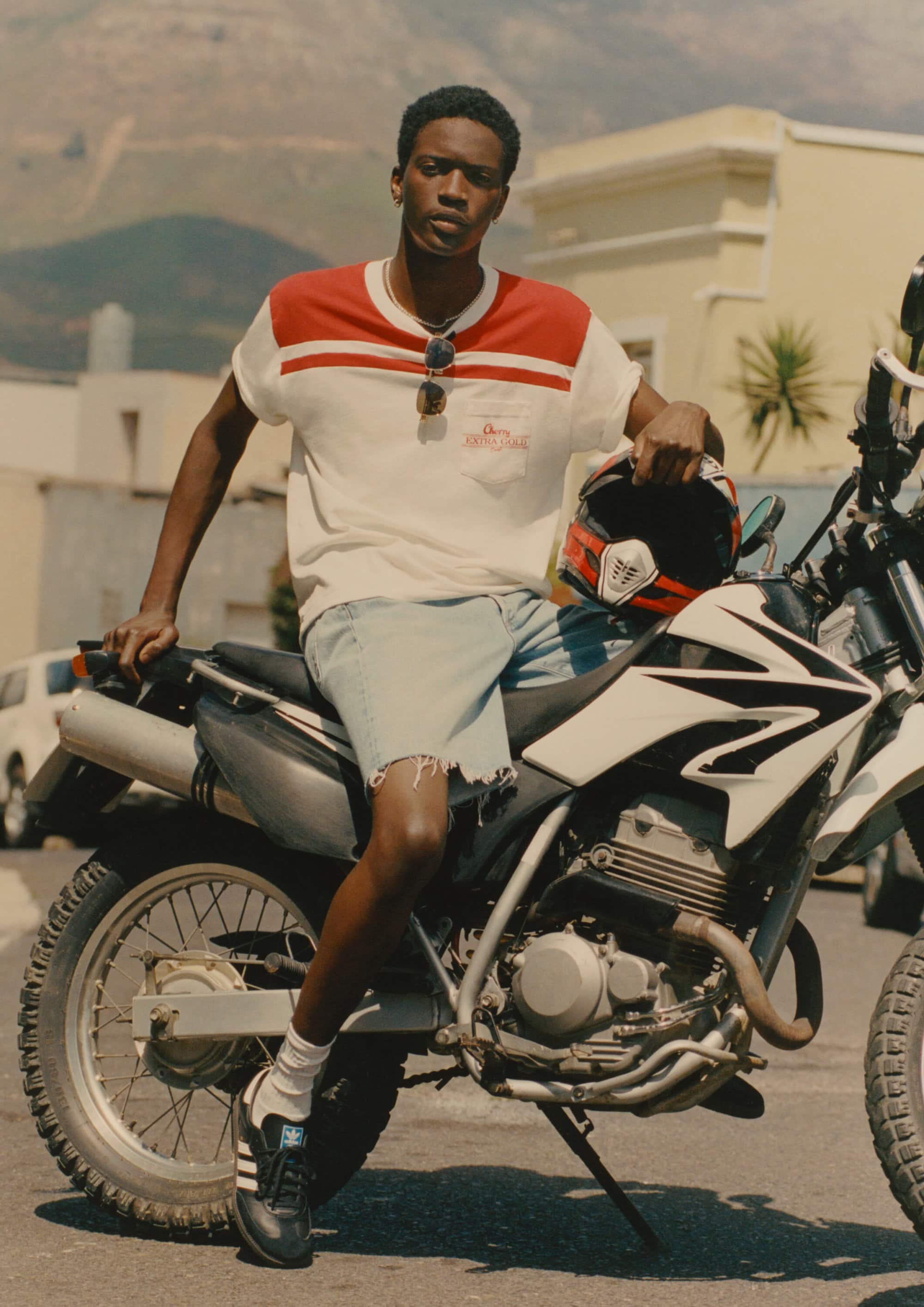 Model Tino Chinyani sitting on a motorcycle holding a helmet for Mr Porter High Summer 2025 campaign