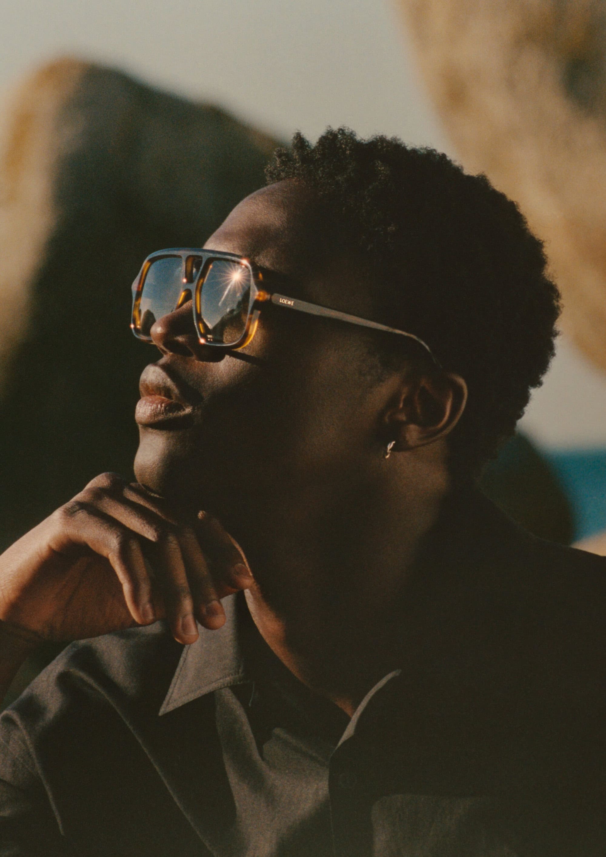 Close-up profile of a black man wearing oversized retro sunglasses and a dark shirt against rocky boulders outdoors