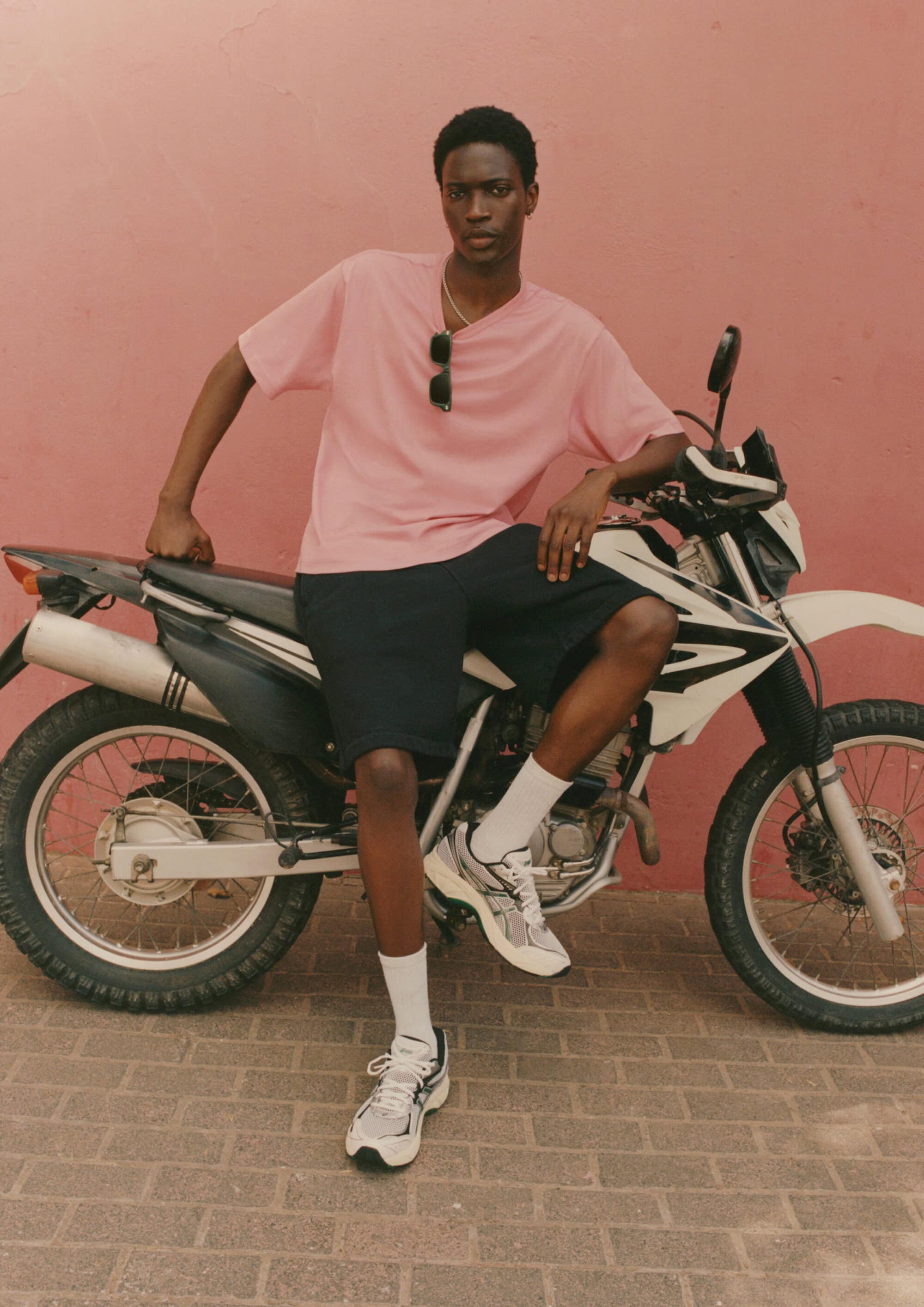 Black man wearing a pink t-shirt and dark shorts sitting on a parked motorcycle against a warm pink wall