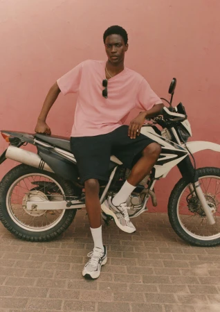 Black man wearing a pink t-shirt and dark shorts sitting on a parked motorcycle against a warm pink wall