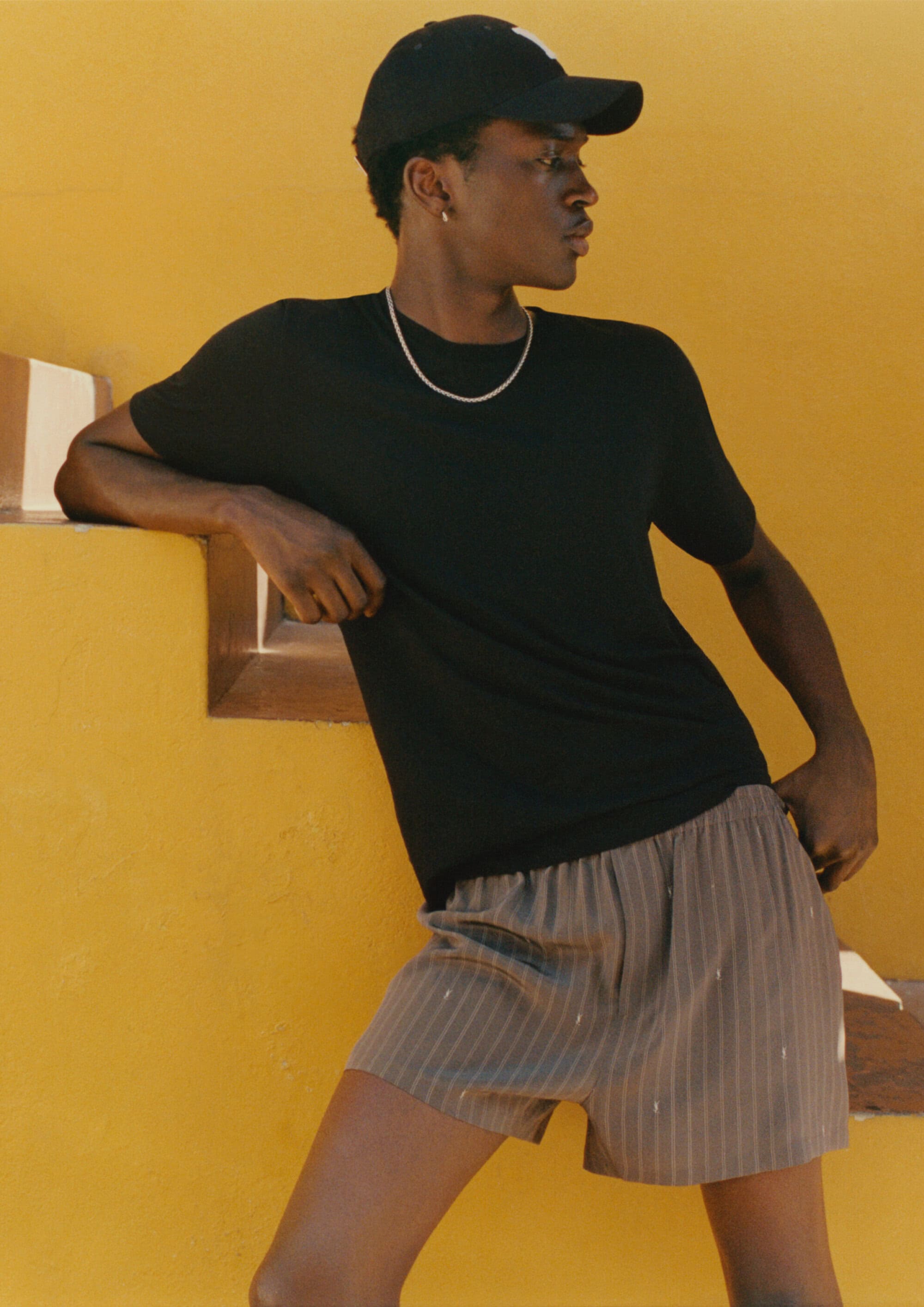 Model wearing a black t-shirt and striped shorts leaning against a yellow wall for Mr Porter