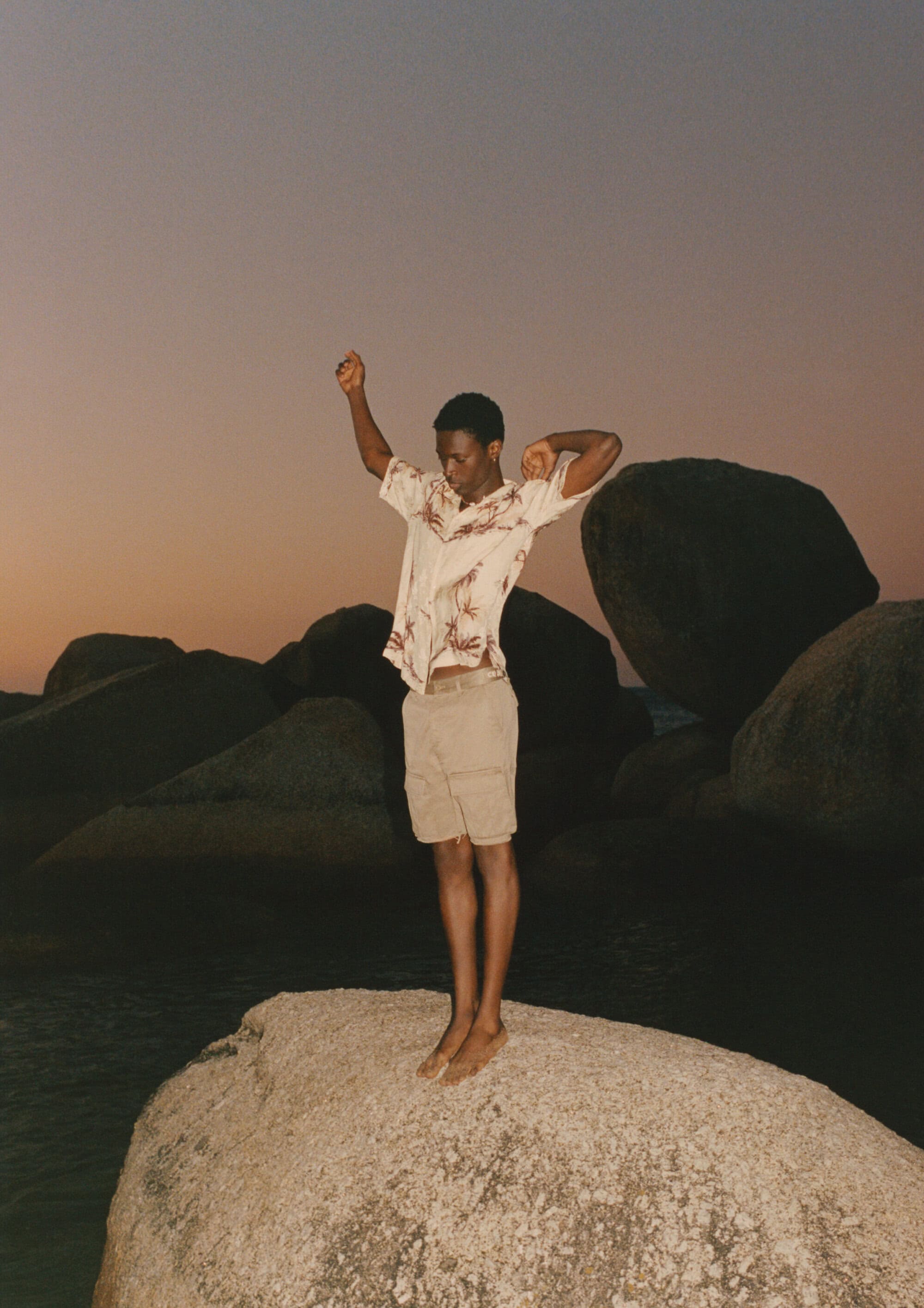 Model Tino Chinyani wearing a patterned short-sleeve shirt standing on a large rock amidst coastal boulders at dusk