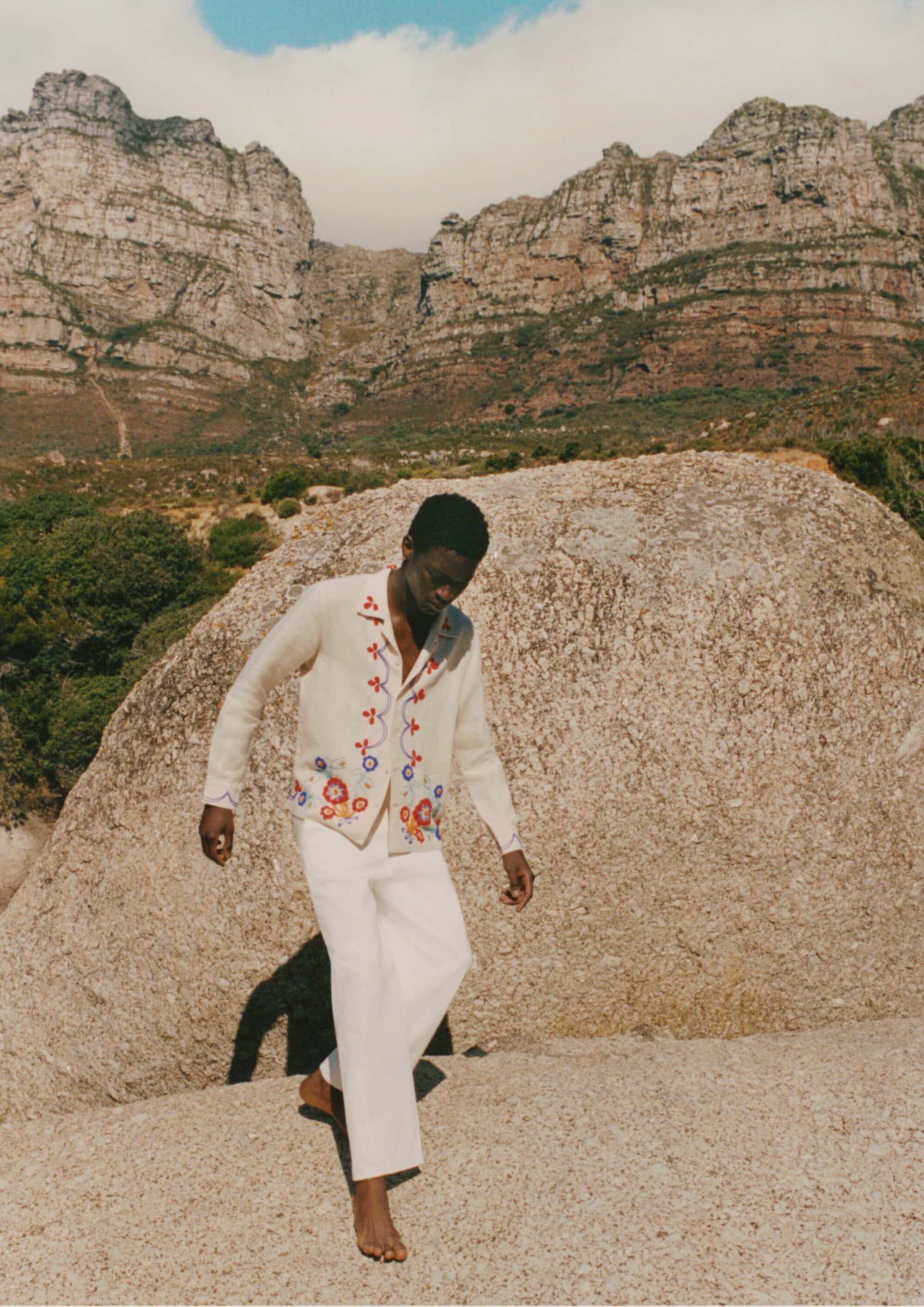 Model Tino Chinyani wearing an embroidered cardigan and white trousers against a rocky mountain landscape.