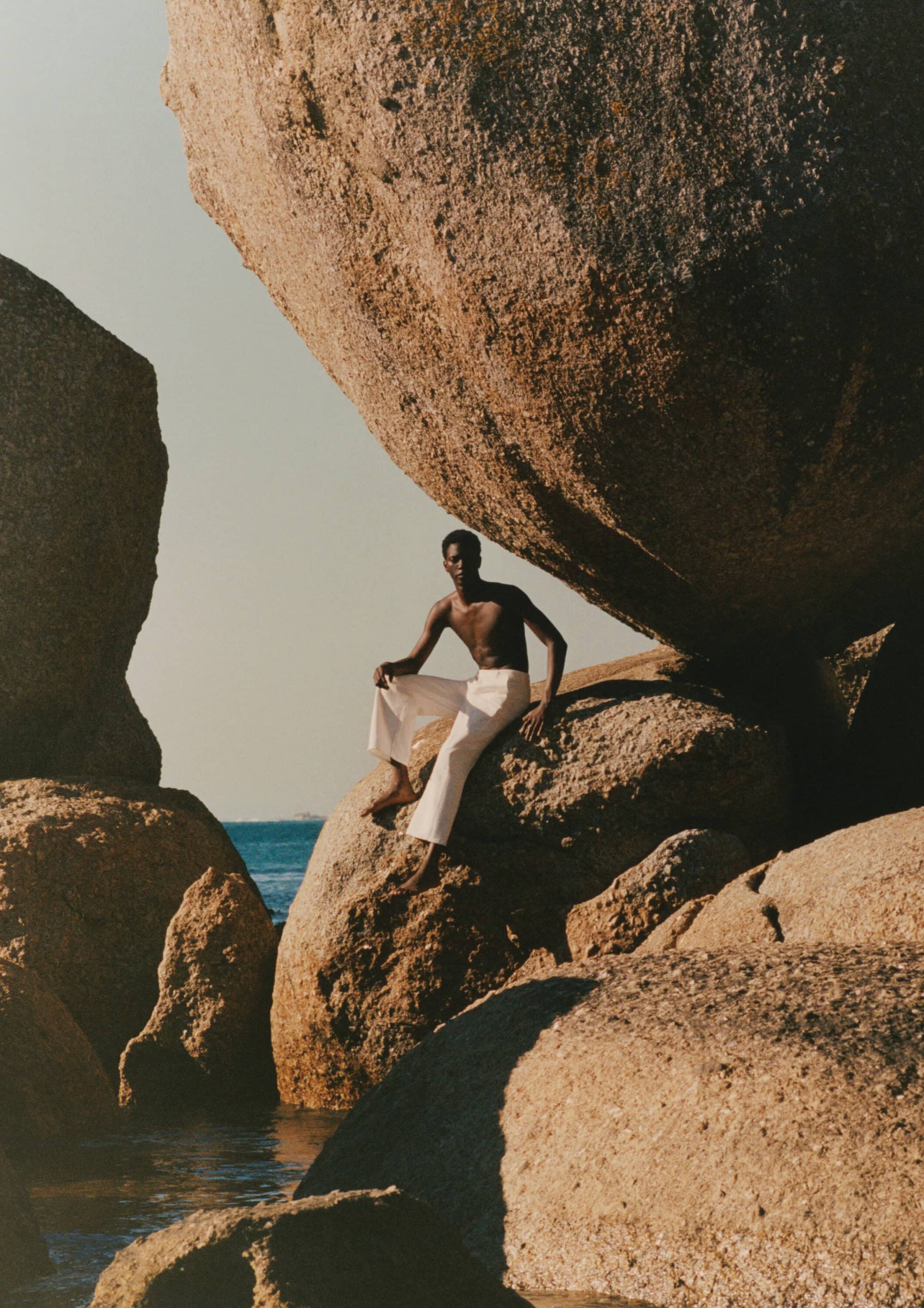 Shirtless man wearing white trousers seated on large boulders by the sea for Mr Porter High Summer 2025 campaign.