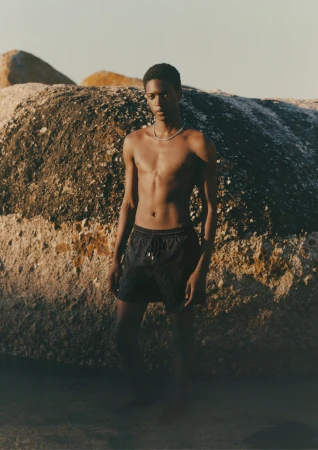 Model Tino Chinyani wearing dark swim shorts standing against large sunlit boulders in a natural outdoor setting.