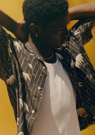 Model Tino Chinyani wearing a patterned short-sleeve shirt over a white tank top against a yellow background.