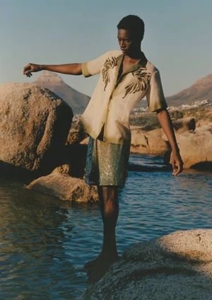 Model balancing on coastal rocks over water wearing an embroidered short sleeve shirt and textured shorts