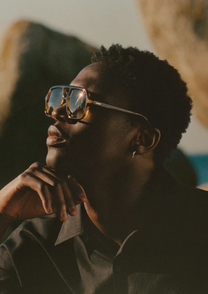 Close-up profile of a black man wearing oversized retro sunglasses and a dark shirt against rocky boulders outdoors