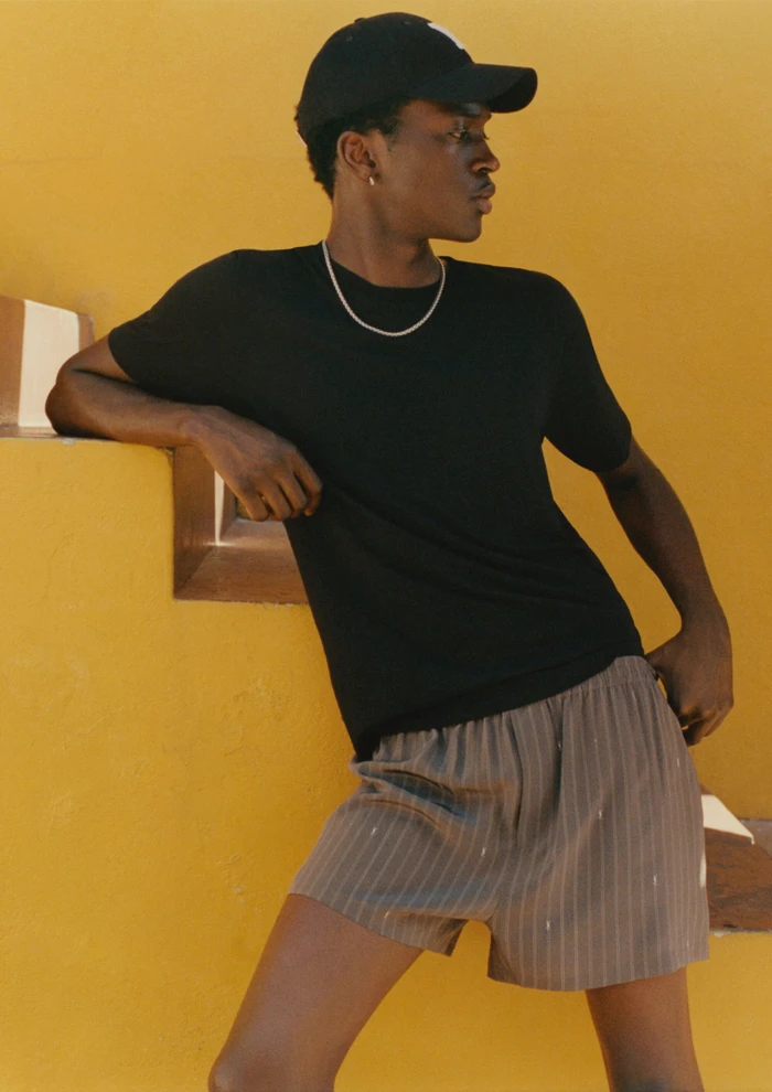 Model wearing a black t-shirt and striped shorts leaning against a yellow wall for Mr Porter