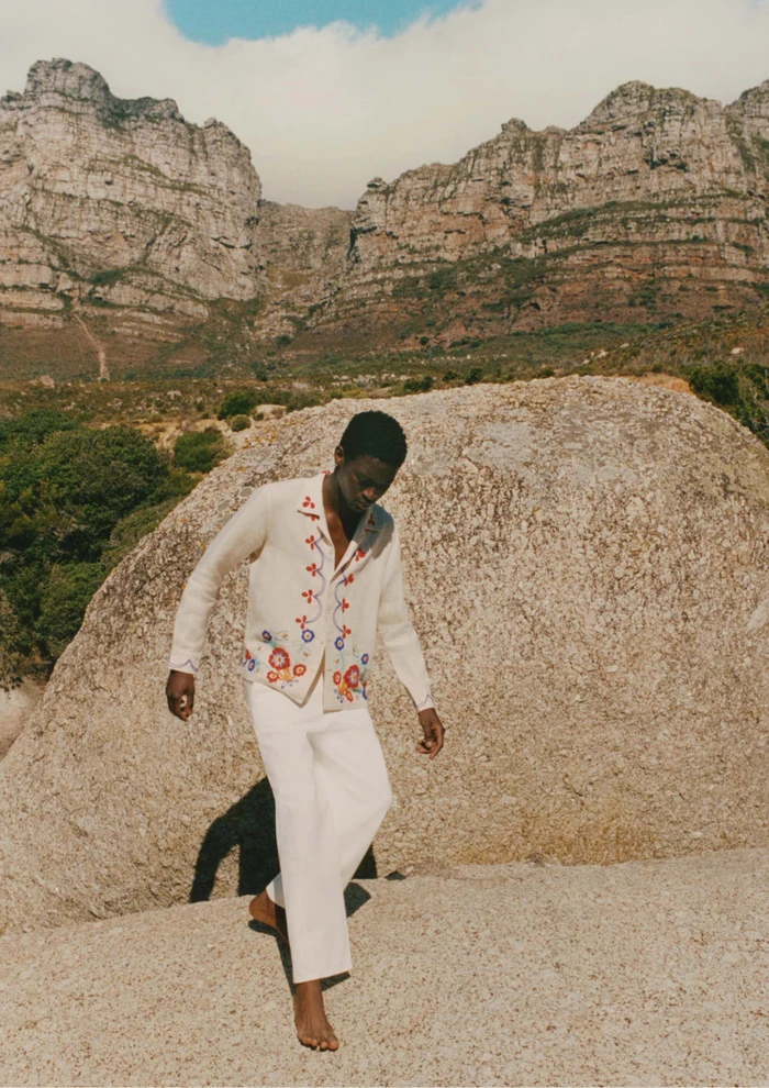 Model Tino Chinyani wearing an embroidered cardigan and white trousers against a rocky mountain landscape.