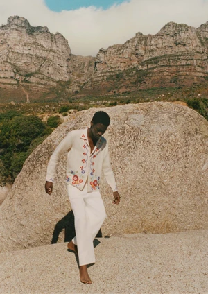 Model Tino Chinyani wearing an embroidered cardigan and white trousers against a rocky mountain landscape.