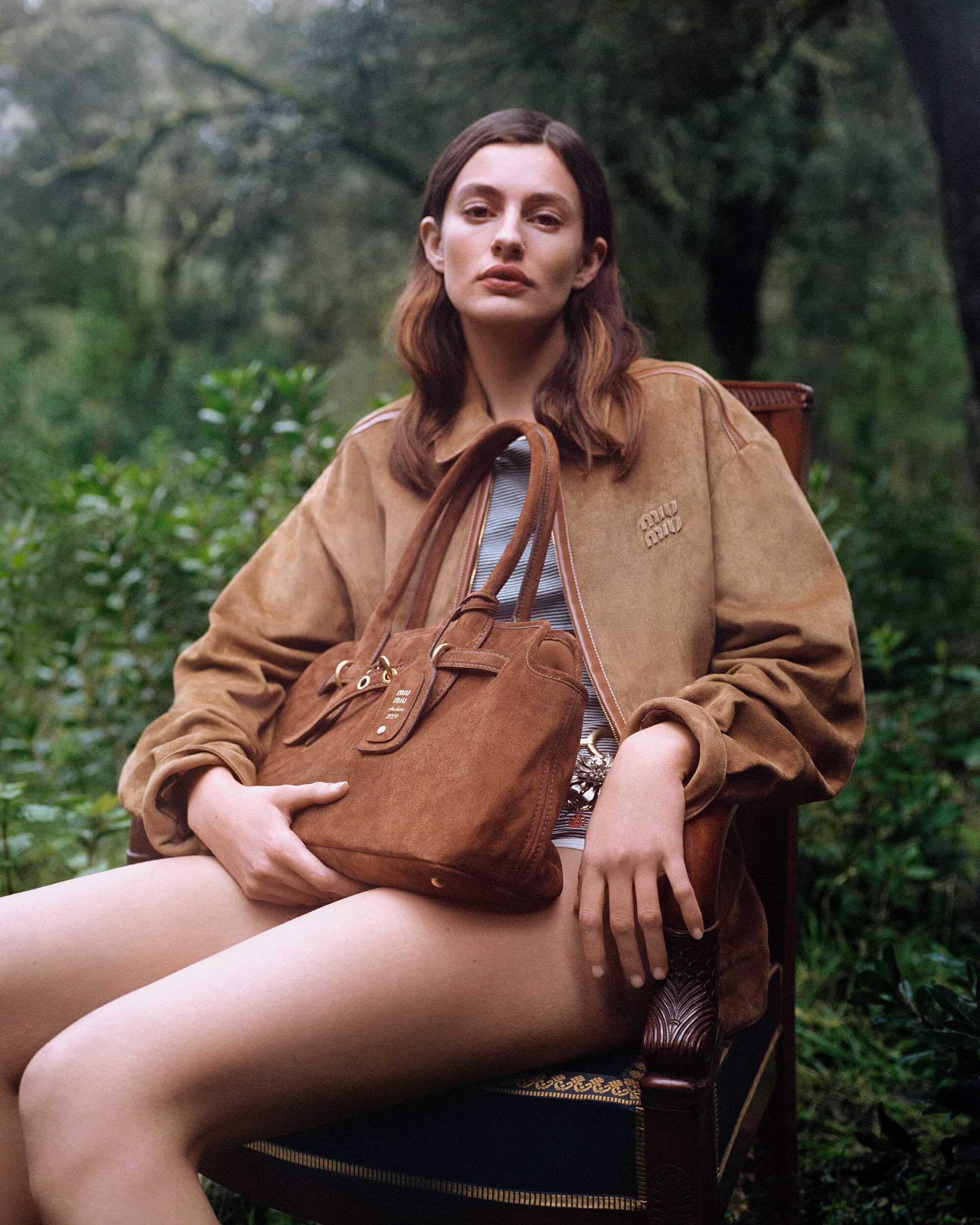 Woman sitting outdoors in a wooden chair wearing a camel suede jacket and holding a matching leather handbag