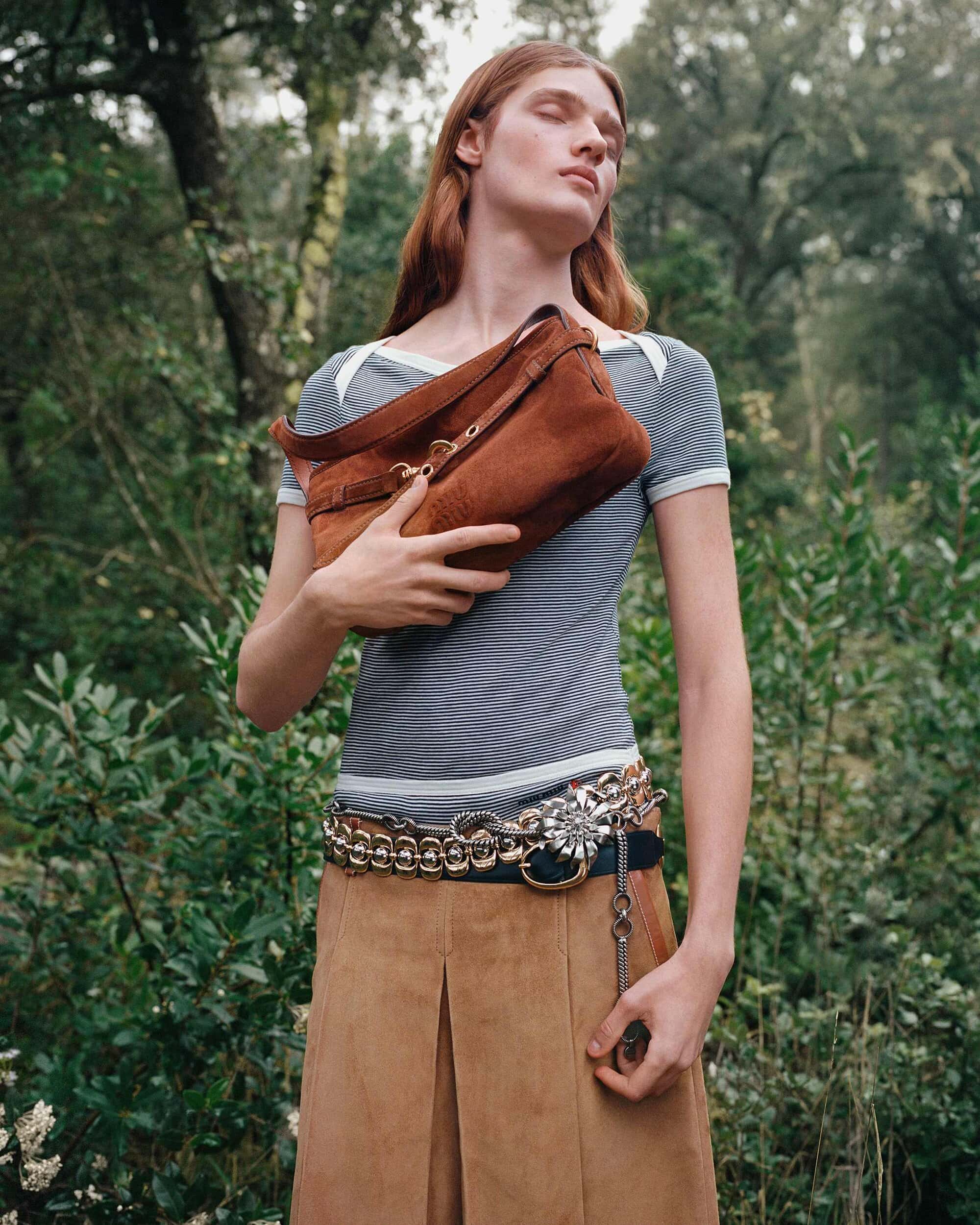 A person with long reddish hair holding a brown suede handbag outdoors amidst trees, wearing a striped top and tan skirt.