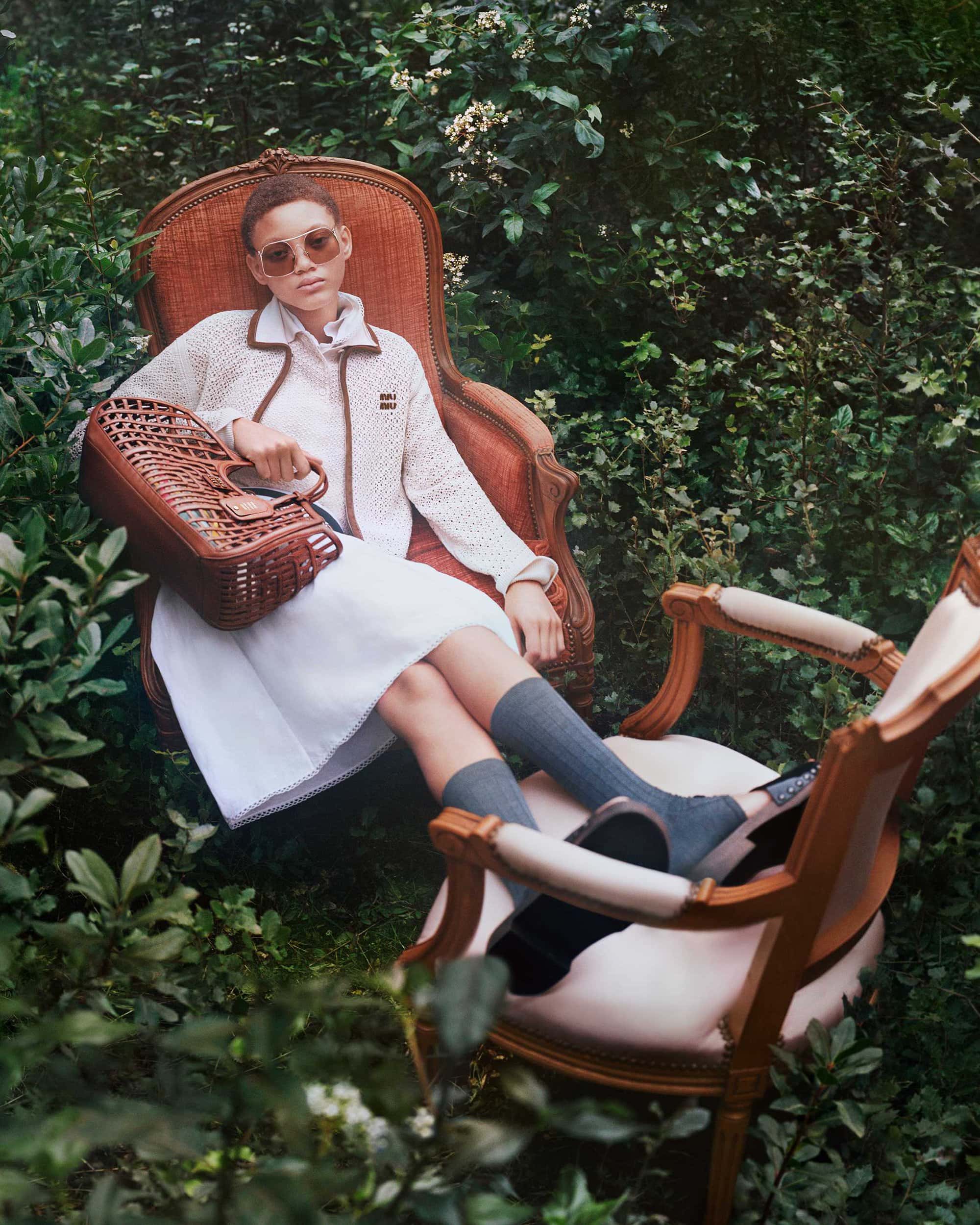 Model wearing sunglasses and white jacket seated outdoors in an armchair holding a brown woven bag for Miu Miu