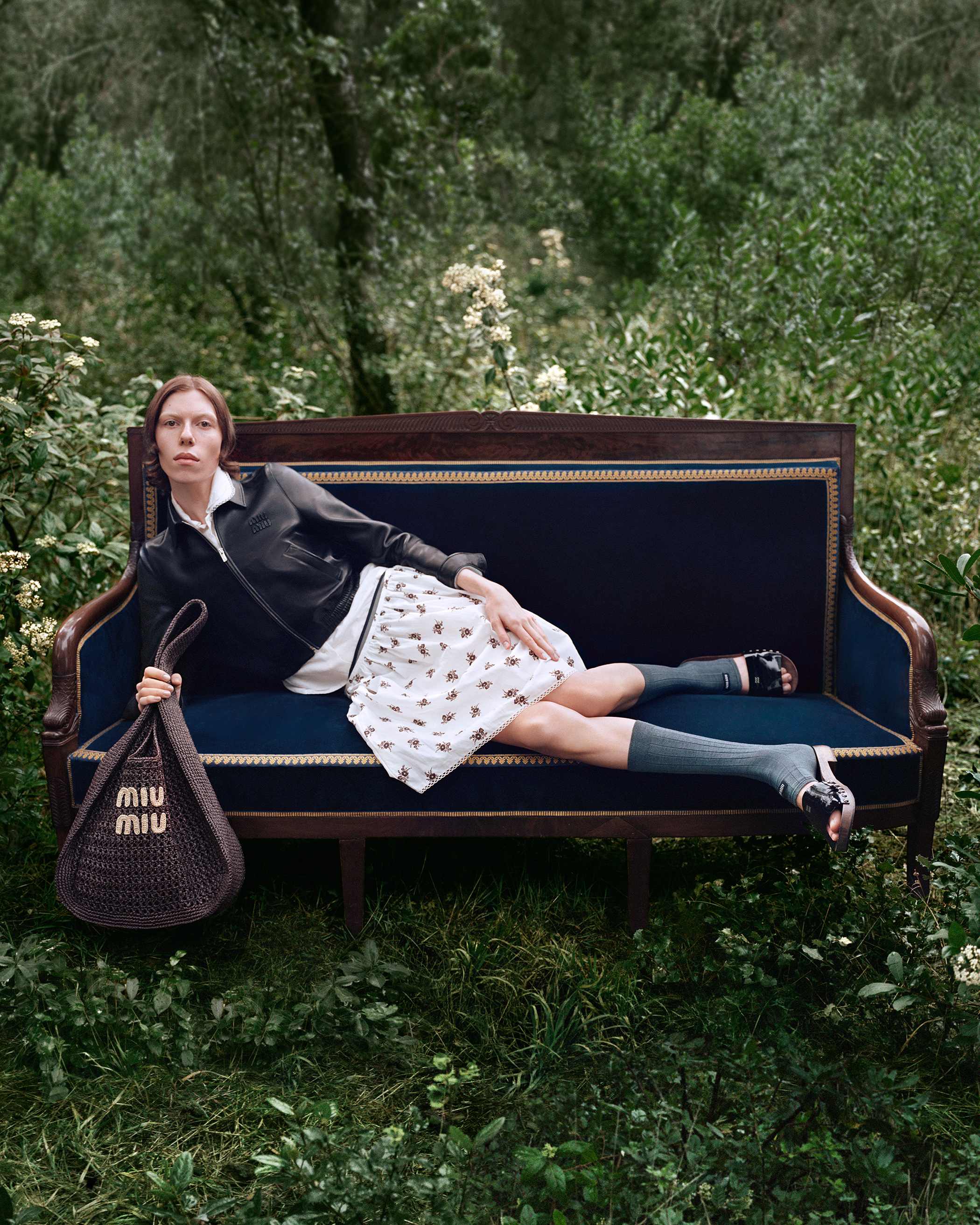 Model reclining on an antique velvet sofa outdoors in a lush forest holding a brown woven Miu Miu bag