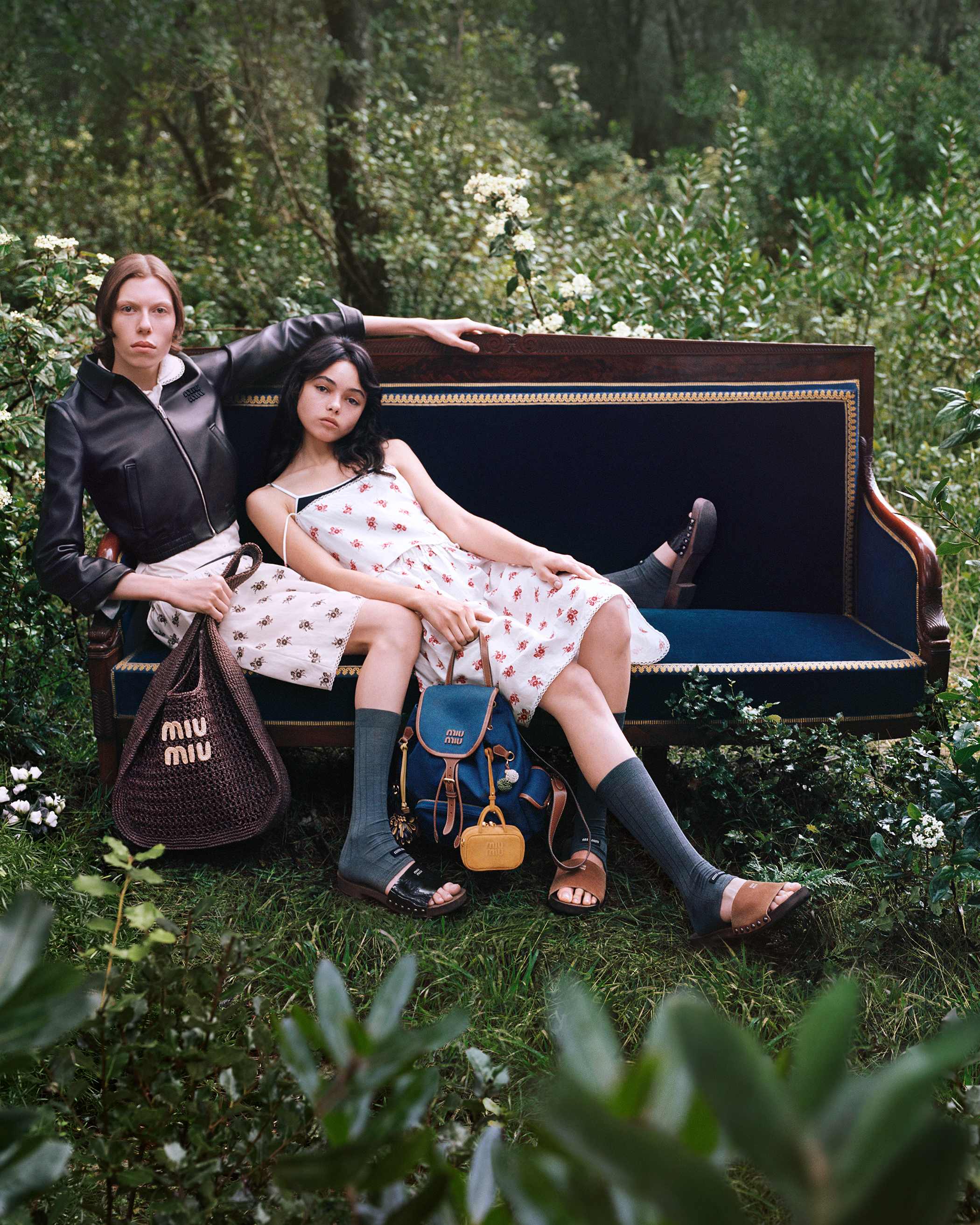 Two models posing on a vintage wooden sofa outdoors in a lush green garden for the Miu Miu L’Eté 2025 ad campaign.