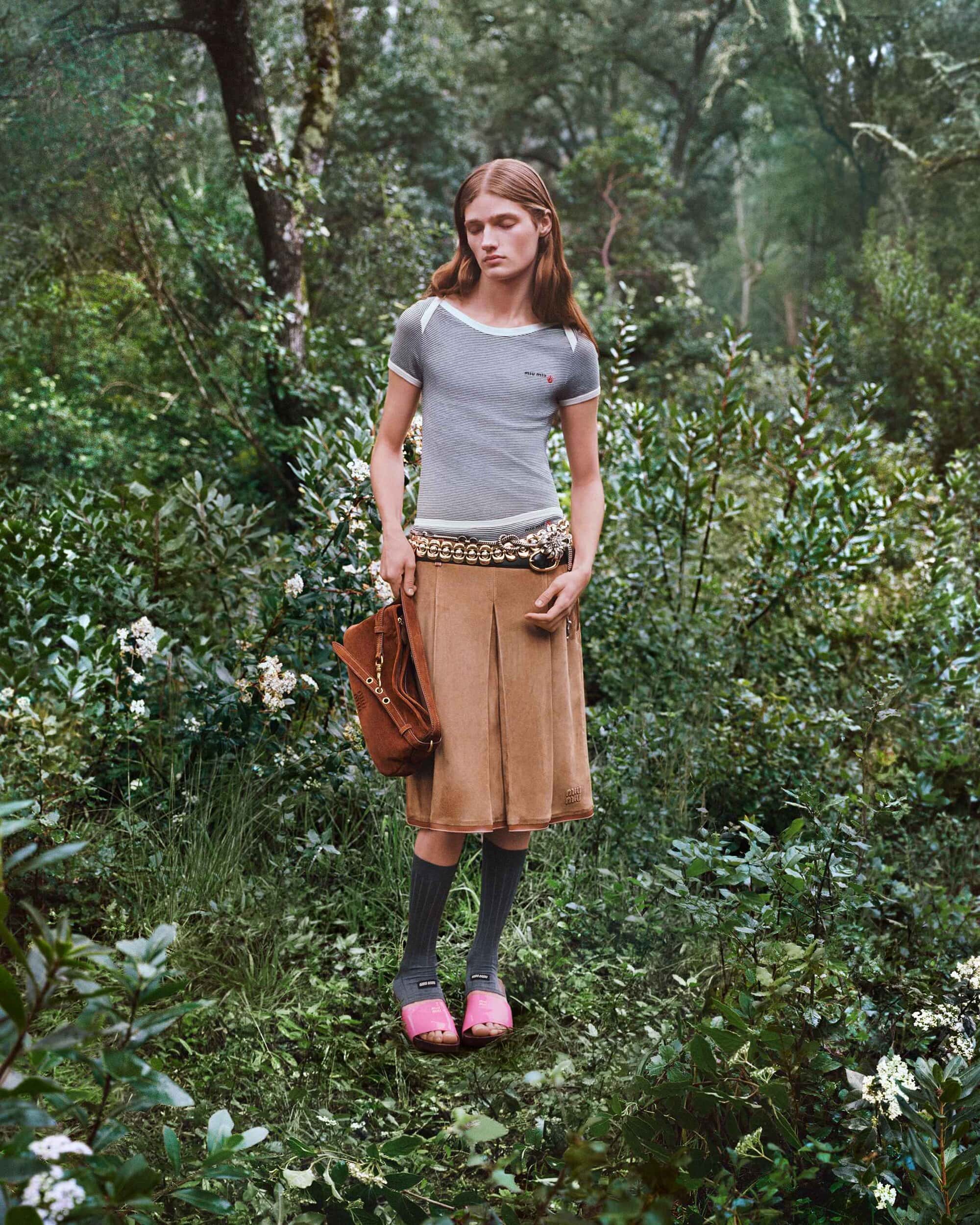 Model standing outdoors in a lush forest wearing a striped t-shirt, brown skirt, high socks, and holding a brown shoulder bag.
