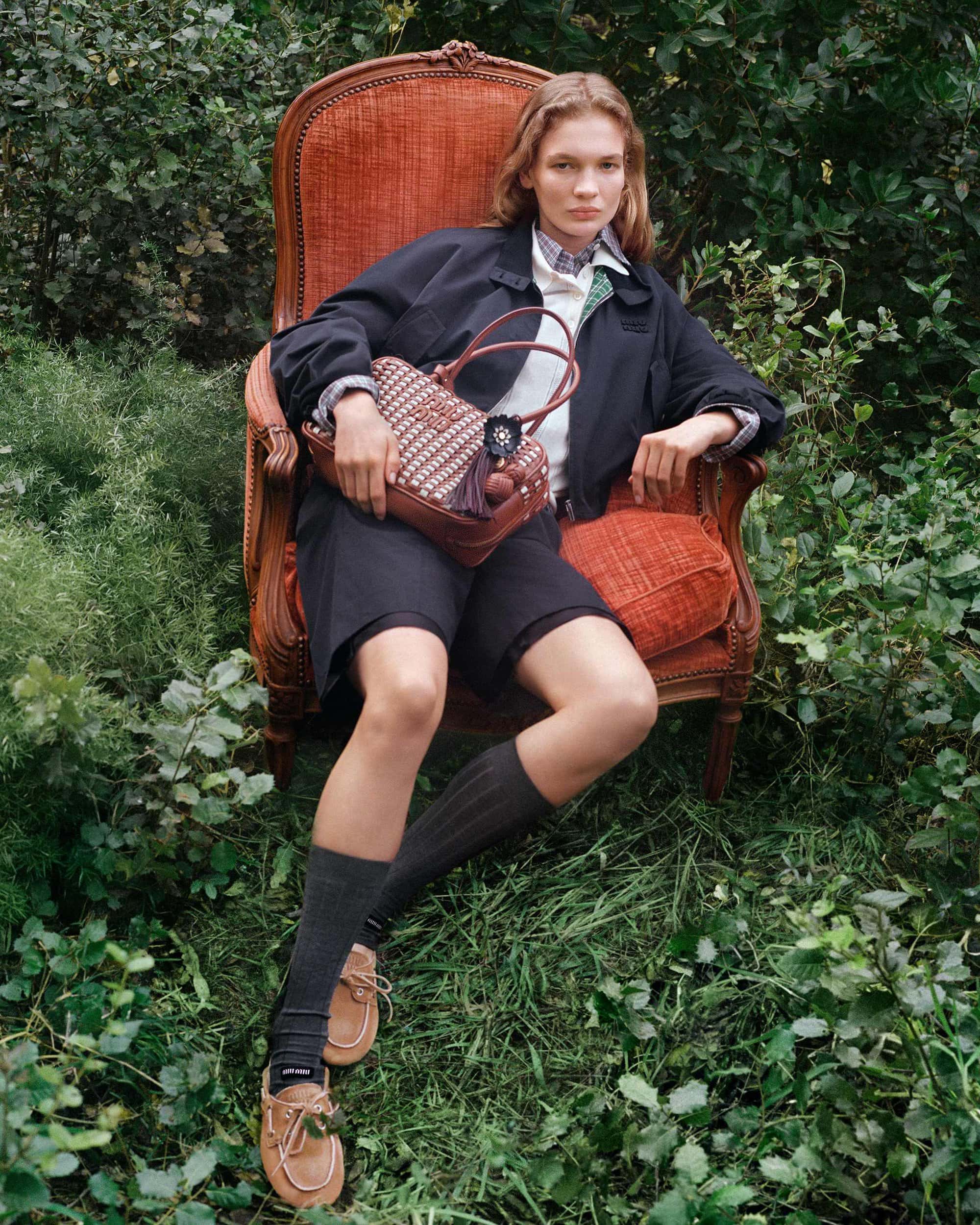 Model seated in an ornate wooden armchair amidst lush greenery holding a brown woven leather handbag