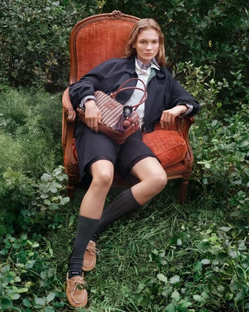 Model seated in an ornate wooden armchair amidst lush greenery holding a brown woven leather handbag