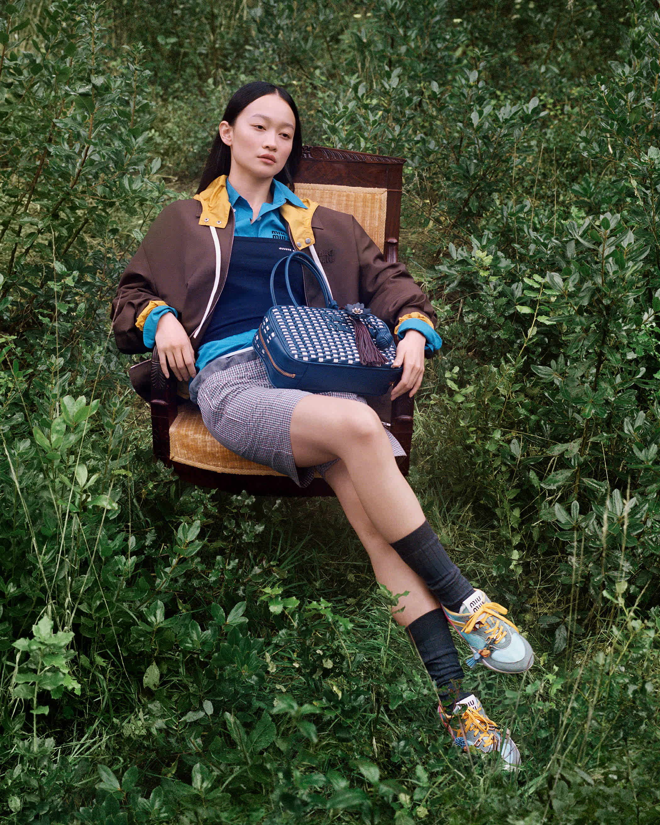 Model seated in an outdoor wooden armchair holding a blue handbag amidst dense green foliage.