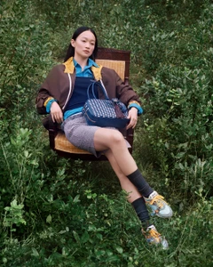 Model seated in an outdoor wooden armchair holding a blue handbag amidst dense green foliage.