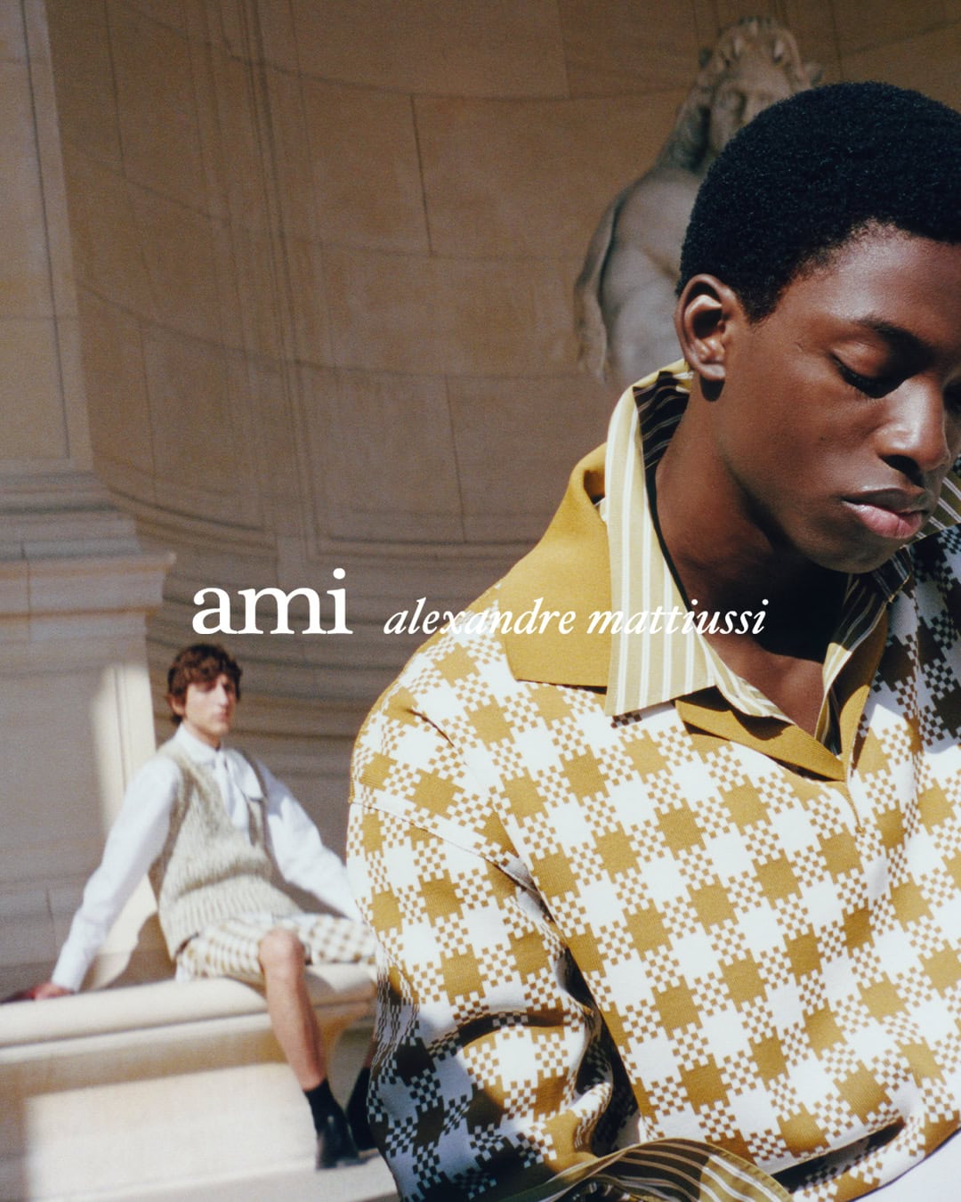 Black man in patterned shirt in the foreground with another man seated in the background against classical stone architecture