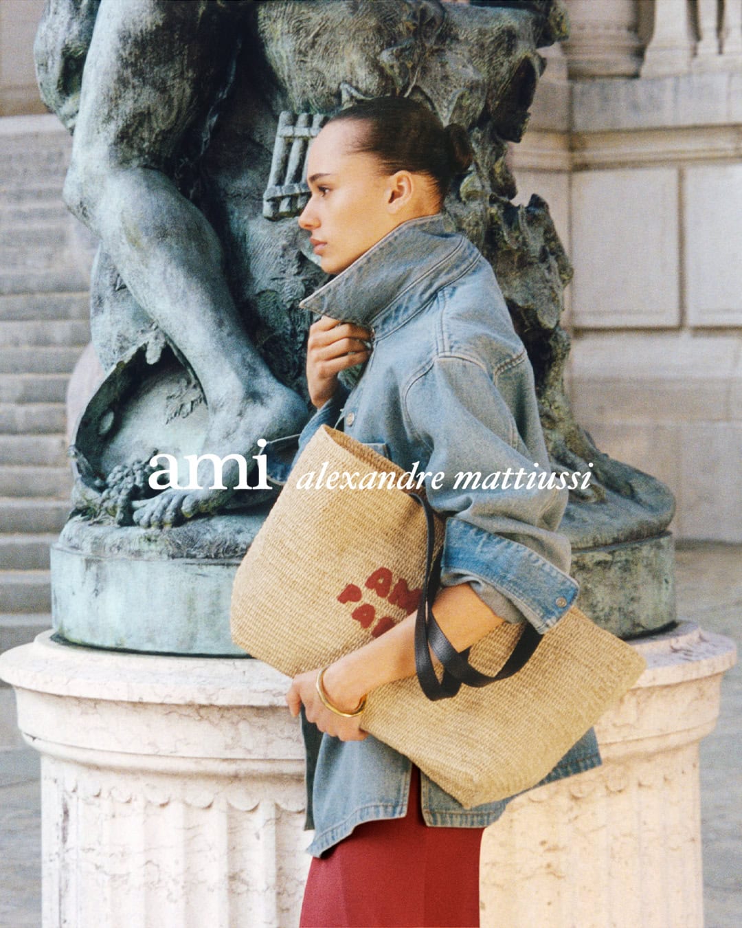 Model in denim jacket holding straw tote bag standing before stone monument for Ami Spring 2025 campaign