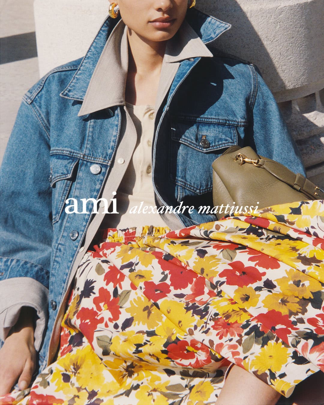Model wearing a blue denim jacket over a beige shirt and red floral skirt holding an olive handbag