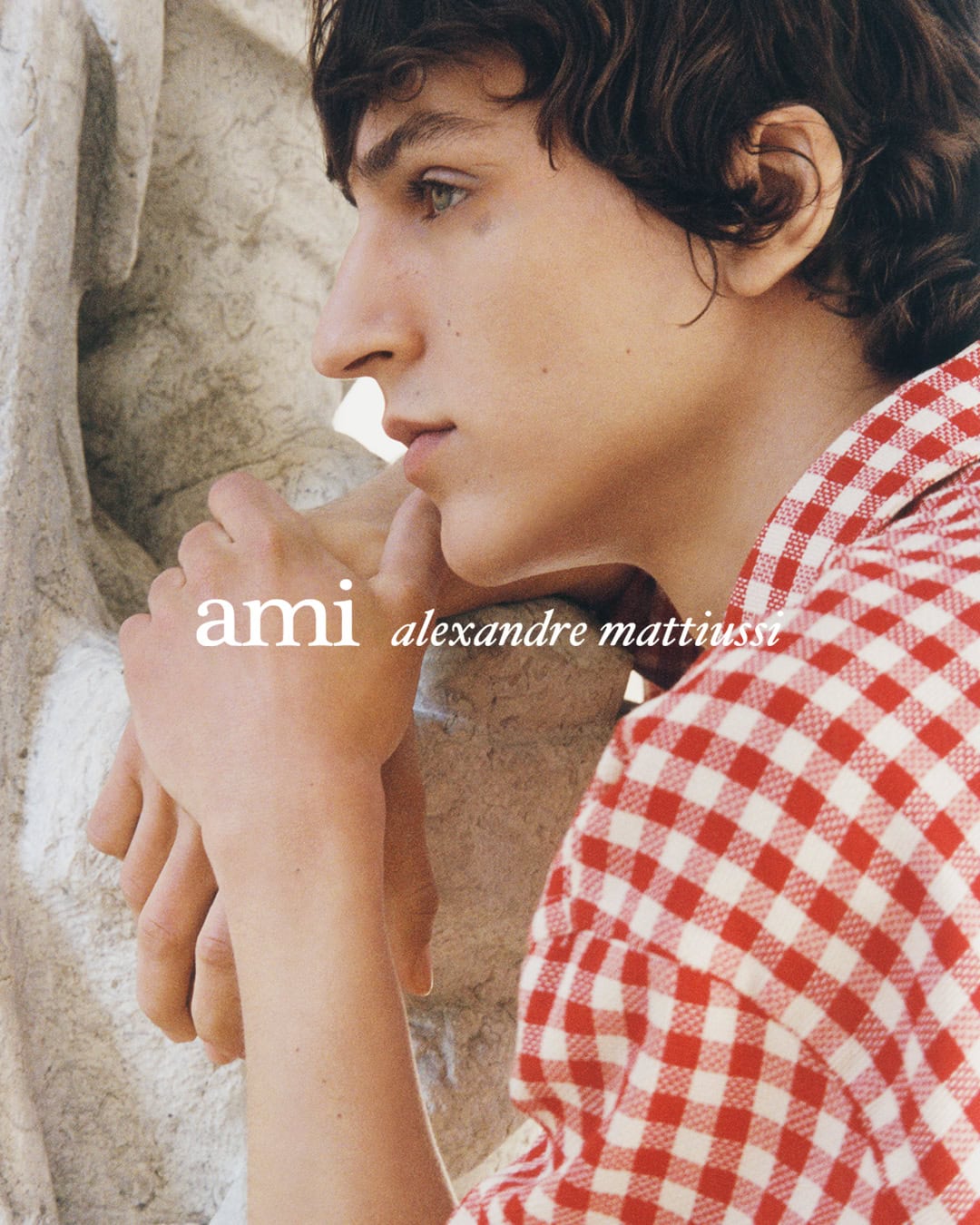 Close-up profile of a young man wearing a red and white checkered shirt leaning against stone architecture.