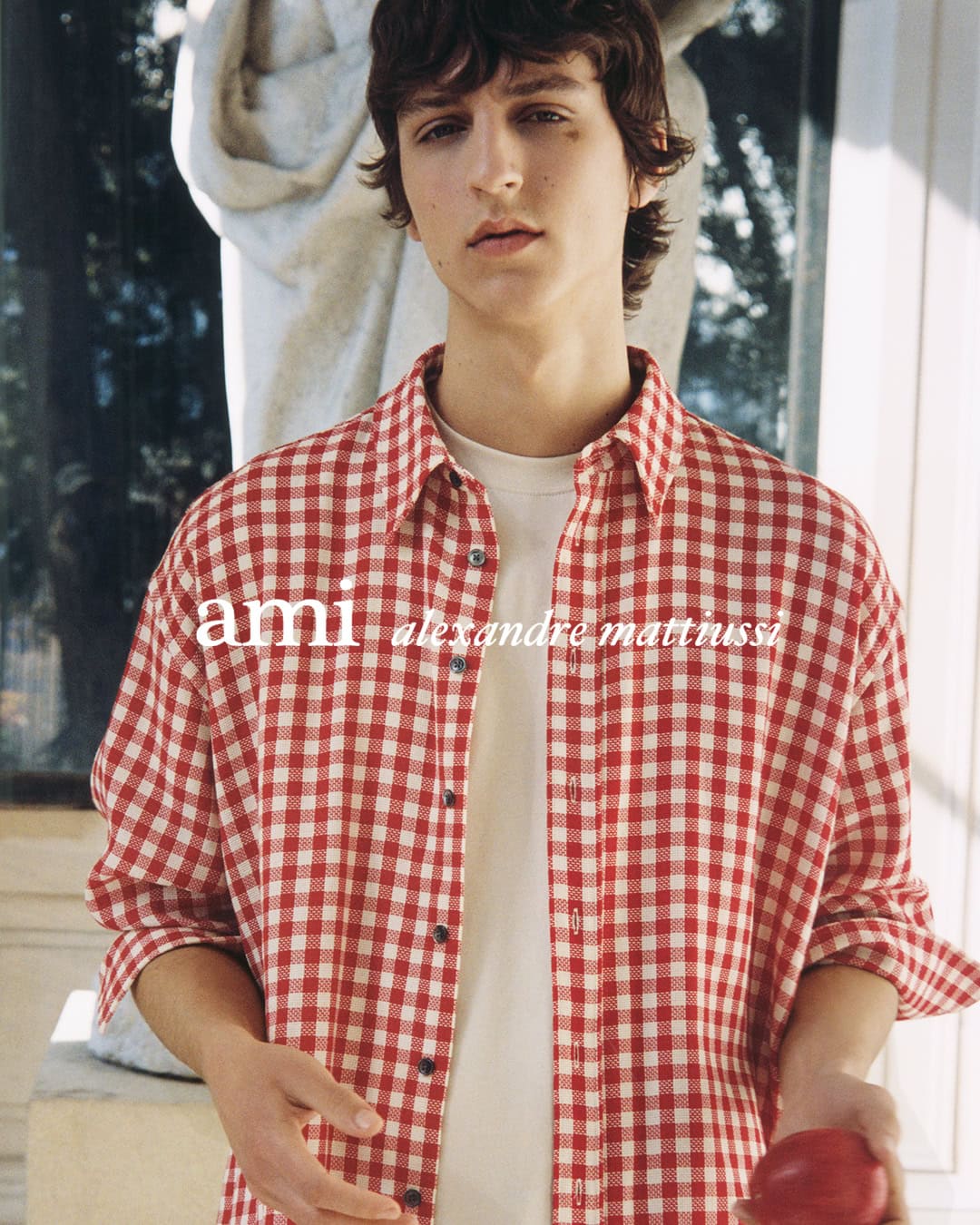 Young man wearing a red and white checkered shirt over a white t-shirt holding a red fruit outdoors in Paris.