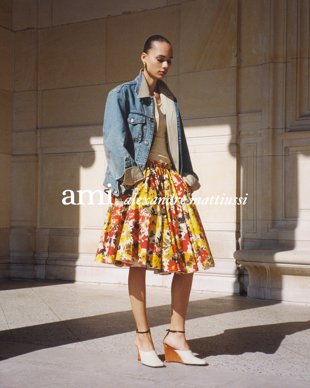 Model standing outdoors wearing an oversized denim jacket over a floral skirt against stone architecture.